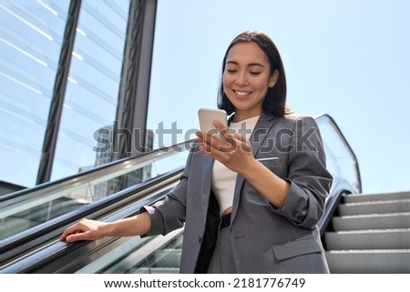 Smiling young adult Asian business woman wearing suit standing on urban escalator using applications on cell phone gadget, reading news on smartphone, browsing fast mobile internet outdoors. Royalty-Free Stock Photo #2181776749