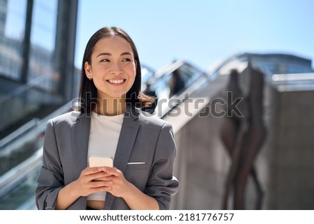 Happy young Asian business woman standing on urban street using cell phone. Smiling lady wearing suit holding smartphone outside advertising modern smart city apps, mobile applications.