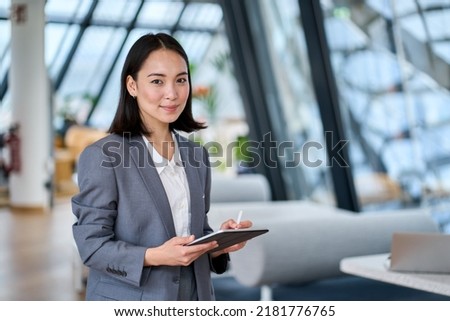 Smiling young Asian business woman leader holding digital tablet standing in office. Professional executive manager or saleswoman using corporate technology looking at camera. Portrait Royalty-Free Stock Photo #2181776765
