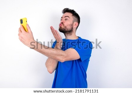 Young caucasian man wearing blue T-shirt over white background blows air kiss at camera of smartphone and takes selfie, sends mwah via online call.