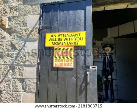 The door with signs on it to a haunted house with a skeleton on the inside waiting. The sign says "Warning Strobe Lights and Loud Music". Another says "An attendant will be with you momentarily".