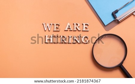 Wooden letters with phrase we are hiring on color background with magnifying glass and clip-board top view. Search worker concept.