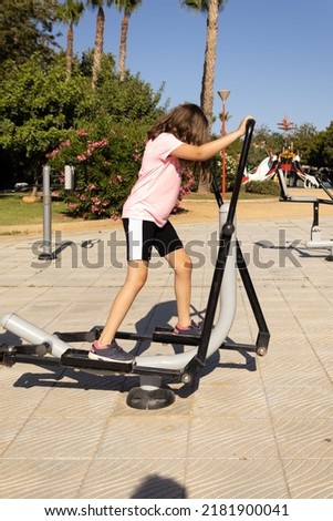 little girl doing sport on a public machine in a park wearing a pink t-shirt and black trousers with a white line, vertical picture