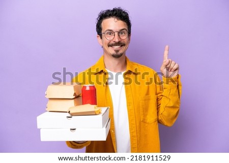 Young caucasian man holding fast food isolated on purple background showing and lifting a finger in sign of the best
