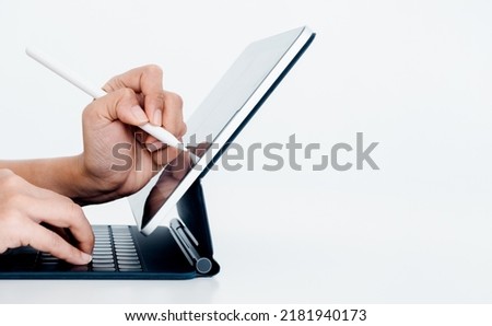 White electronic pen or pencil in person's hand, drawing on digital tablet screen while typing on keyboard computer isolated on white background with copy space, side view. Work with technology.
