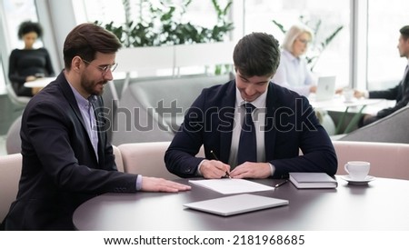 Happy young Caucasian male employee put signature on document close deal with employer or boss in office. Young businessmen or partners sign contract paperwork make business agreement at meeting.