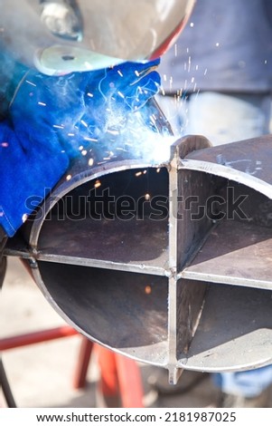 A professional welder in a protective suit and mask produces the connection of pipes with the help of a welding machine on the construction site. Royalty-Free Stock Photo #2181987025