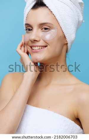 a beautiful woman stands on a blue background, with a towel on her head after a shower and patches pasted on her face, looks away holding her hand near her face and smiles happily