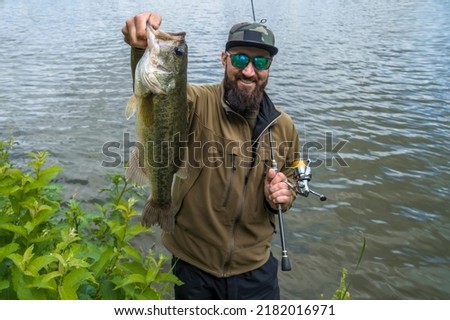 Bass fishing. Large bass fish in hands of pleased bearded fisherman with tackle. Largemouth perch at pond Royalty-Free Stock Photo #2182016971