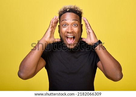 Black fan in a black T-shirt and smartwatch shouts loudly on a yellow background. The joyful African opened his mouth wide and bulged his eyes loudly delivering wonderful news Royalty-Free Stock Photo #2182066309