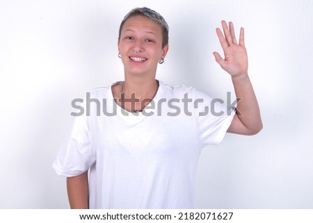 young woman with short hair wearing white t-shirt over white background waiving saying hello or goodbye happy and smiling, friendly welcome gesture.
