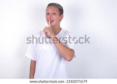 Smiling young woman with short hair wearing white t-shirt over white background makes shush gesture, holds fore finger over lips hides secret. Be mute, please.