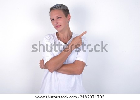 young woman with short hair wearing white t-shirt over white background smiling broadly at camera, pointing fingers away, showing something interesting and exciting.
