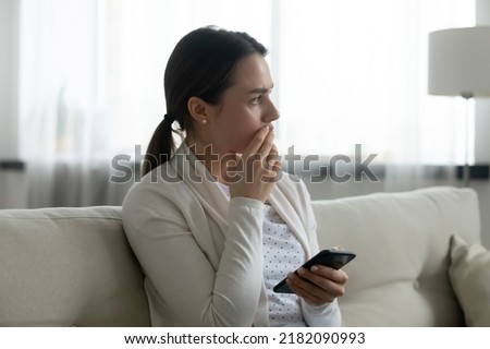 Pensive stressed young Caucasian woman sit on sofa at home using cellphone look in distance pondering of problem. Anxious millennial girl feel distressed frustrated with message or text on smartphone. Royalty-Free Stock Photo #2182090993