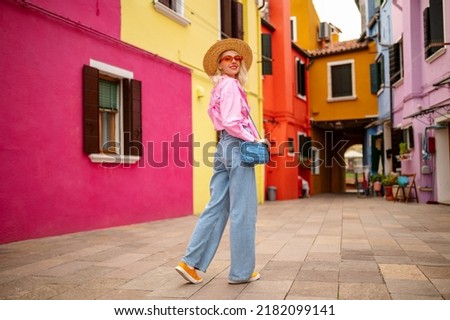 Happy smiling female traveler wearing stylish hat, glasses, pink shirt, wide leg trousers, walking, posing near colorful houses in street. Travel, tourism, vacation, fashion, lifestyle conception Royalty-Free Stock Photo #2182099141