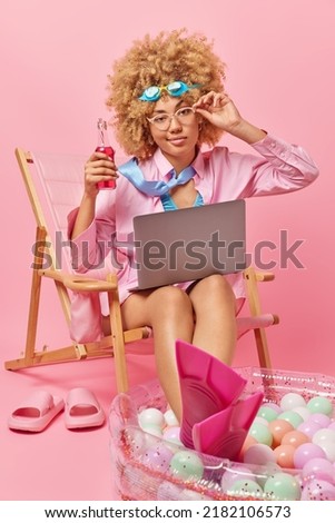 Photo of serious woman dreams about summer vacation focused at camera with attentive gaze uses laptop computer drinks energetic beverage keeps hand on rim of spectacles sits on lounge chair.