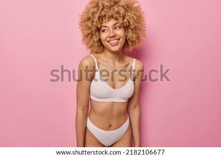 Horizontal shot of cheerful woman with curly hair looks away gladfully dressed in cropped top and panties demonstrates perfect figure healthy skin isolated over pink background. Wellness and body