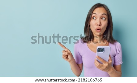 Amazed Asian woman with dark hair points aside shows advertisement uses mobile phone enjoys online communication dressed in casual purple t shirt isolated over blue background. Technology and reaction Royalty-Free Stock Photo #2182106765