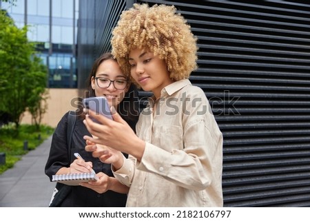 Outdoor shot of two mixed race student girls watch something via mobile phone write down notes in spiral notebook cooperate for completing common project browse social media discuss media files