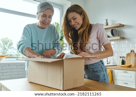Excited senior woman and her adult daughter unpacking box while standing at the domestic kitchen Royalty-Free Stock Photo #2182111265