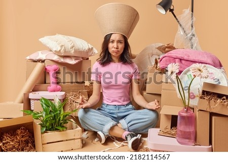 Unhappy Asian woman meditates sits in lotus pose tries to relax poses around packages full of property moves in new place for living isolated over beige background. Moving and mortgage concept Royalty-Free Stock Photo #2182114567