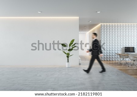 Side view of young businessman walking in modern concrete and wooden coworking office interior with empty mock up place on wall, furniture and equipment. CEO and executive concept Royalty-Free Stock Photo #2182119801