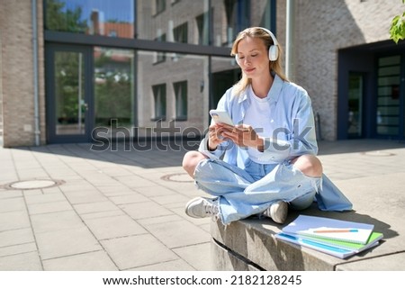 Girl university student wearing headphones using smartphone app sitting on stairs outdoors online learning, remote studying virtual class, watching webinar distance course or listening podcast.