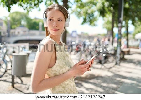 Young pretty woman model standing on summer city street wearing headphones listening to music or podcast audio in mobile streaming service app holding phone using smartphone outdoors.