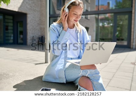 Pretty girl university student wearing headphones using laptop computer sitting on stairs outdoors in campus online learning, remote studying class, watching educational webinar course concept. Royalty-Free Stock Photo #2182128257