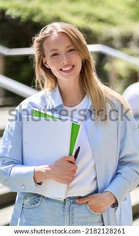 Smiling pretty happy blond girl university or college student holding notebooks looking at camera standing outside campus. Vertical portrait outdoors, education program courses and classes ads.