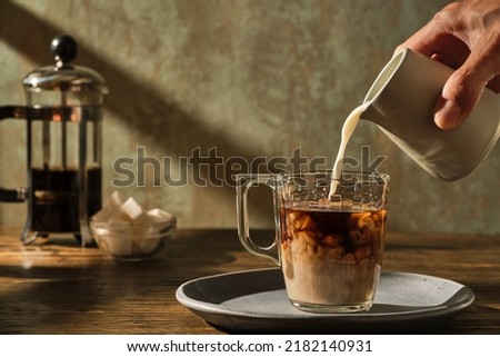 pouring milk into the coffee with a jug with coffee pot and sugar cubes in the background illuminated with warm light from a window Royalty-Free Stock Photo #2182140931