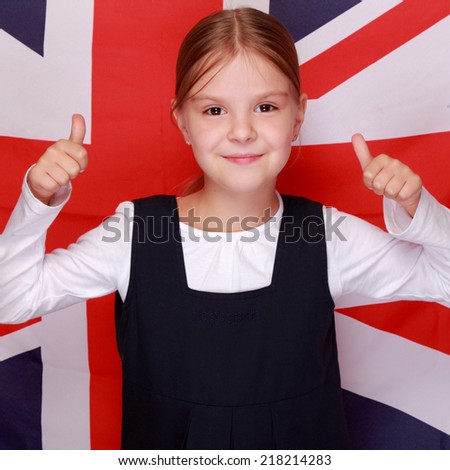 Adorable little girl with a sweet smile in school uniform holding a thumb up on background of the flag of Great Britain/British schoolgirl smiling at the background of the flag of England