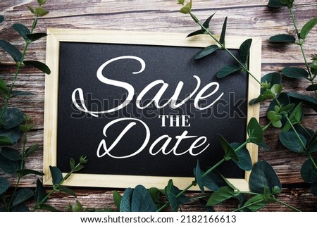 Save the Date typography text written on wooden blackboard with alarm clock and green eucalyptus decoration on wooden background