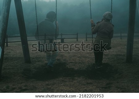 Concept of afterlife and memory for parents or lost love. Back view of woman sitting on a swing with ghost man near her outdoor in the park with fog. . Dead friend or husband concept. Life and death Royalty-Free Stock Photo #2182201953