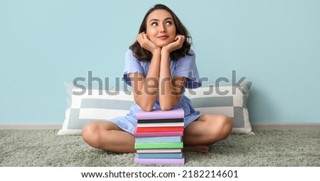 Beautiful thoughtful young woman with many books sitting near light blue wall