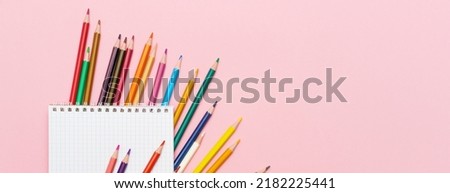 Colored pencils for drawing and a notebook in a cage on a pink background. Back to school concept