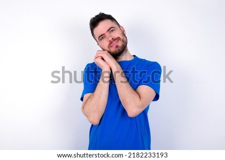 Charming serious Young caucasian man wearing blue T-shirt over white background keeps hands near face smiles tenderly at camera
