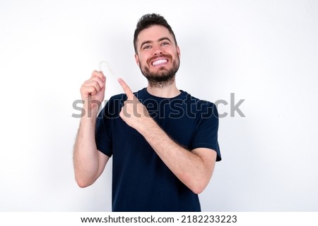 Young caucasian man wearing black T-shirt over white background holding an invisible aligner and pointing at it. Dental healthcare and confidence concept.