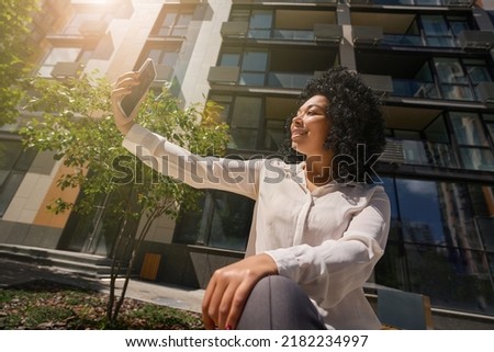 Modern multiracial woman relaxing on bench and taking selfie