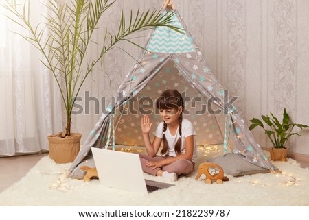 Portrait of cute little girl sitting in peetee tent and having video call or broadcasting livestream, looking at notebook screen, waving hand, saying bye or hello.