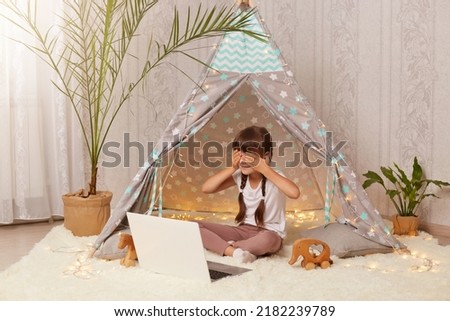 Indoor shot of little preschooler girl sitting in wigwam in front of notebook, having video call, covering eyes with palm, hiding herself, playing in new peetee tent.