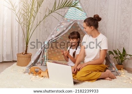 Portrait of dark haired female and her daughter wearing white t shirts sitting in wigwam in front of laptop, child using smart phone, mother looking at kid, family spending time together.