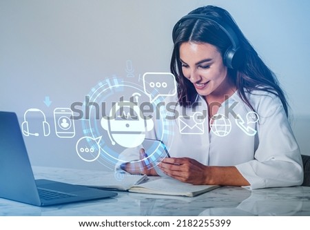 Businesswoman in headphones smiling, using phone, digital chat bot hud hologram. Office table with laptop and notebook. Concept of helpdesk Royalty-Free Stock Photo #2182255399