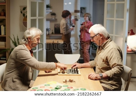 Two friendly senior men sitting by table in living room and playing chess on background of their wives having wine and talking in the kitchen