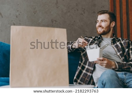 Young fun man he wears brown shirt eat Chinese food cuisine in takeaway carton container box sit on sofa in own living room stay home indoor flat on weekends. Delivery service from shop or restaurant