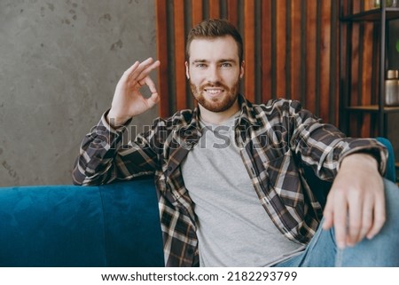 Young satisfied cheerful fun cool man 20s he wears brown shirt showing okay ok gesture sitting on blue sofa in own living room apartment stay home indoor flat on weekends. People lifestyle concept.