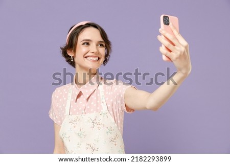 Fun young housewife housekeeper chef cook baker woman in pink apron doing selfie shot on mobile phone post photo on social network isolated on pastel violet background studio. Cooking food concept.