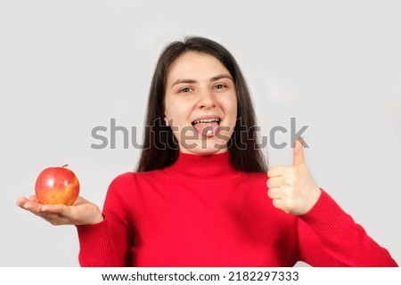 A woman with a pill on her tongue holds a red apple on a white background showing thumbs up