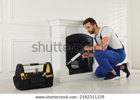 Professional technician sealing electric fireplace with caulk near white wall in room Royalty-Free Stock Photo #2182311339