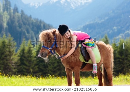 Kids riding pony in the Alps mountains. Family spring vacation on horse ranch in Austria, Tirol. Children ride horses. Kid taking care of animal. Child and pet. Little girl in blooming meadow. Royalty-Free Stock Photo #2182315855